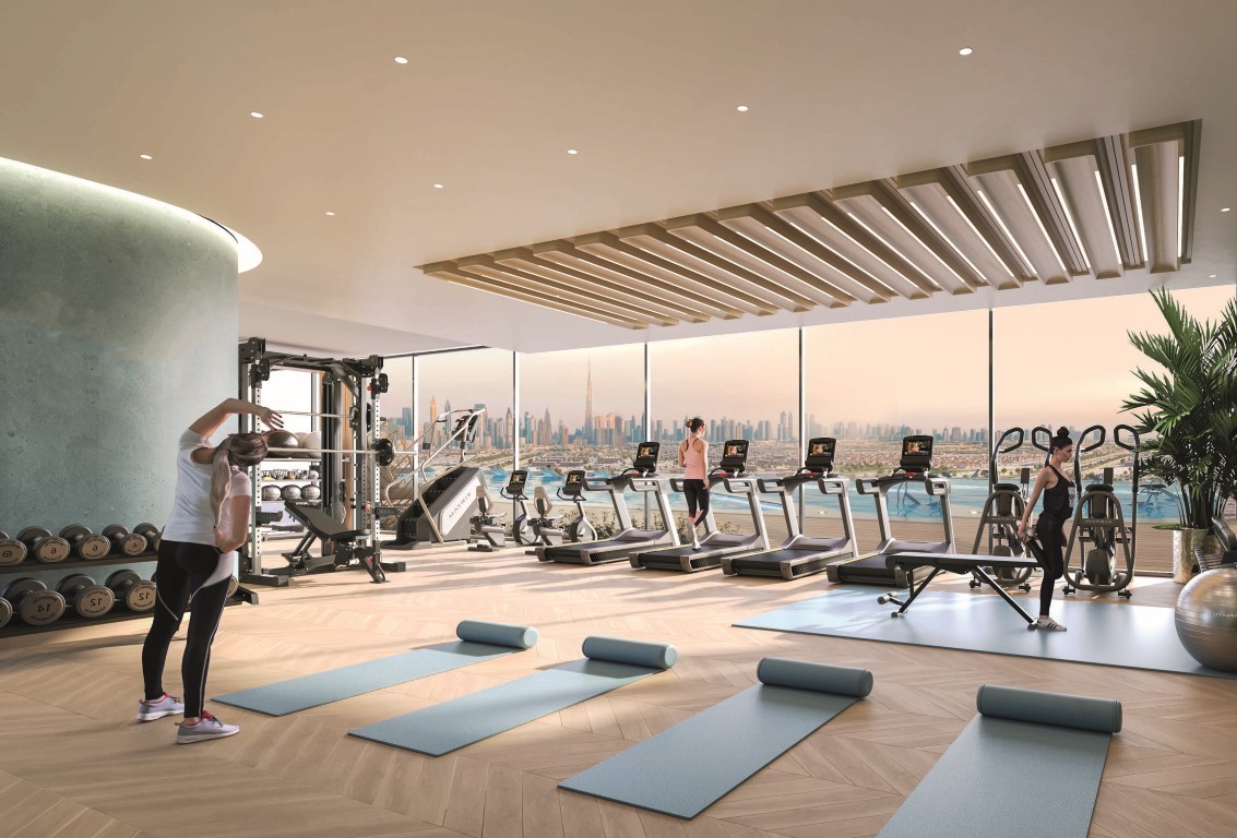 Modern gym with large windows showcasing a city skyline, perfect for residents investing in Dubai Real Estate. Several treadmills, exercise machines, free weights, and three blue yoga mats are set on a wooden floor. Two people stretch as others use the cardio equipment.