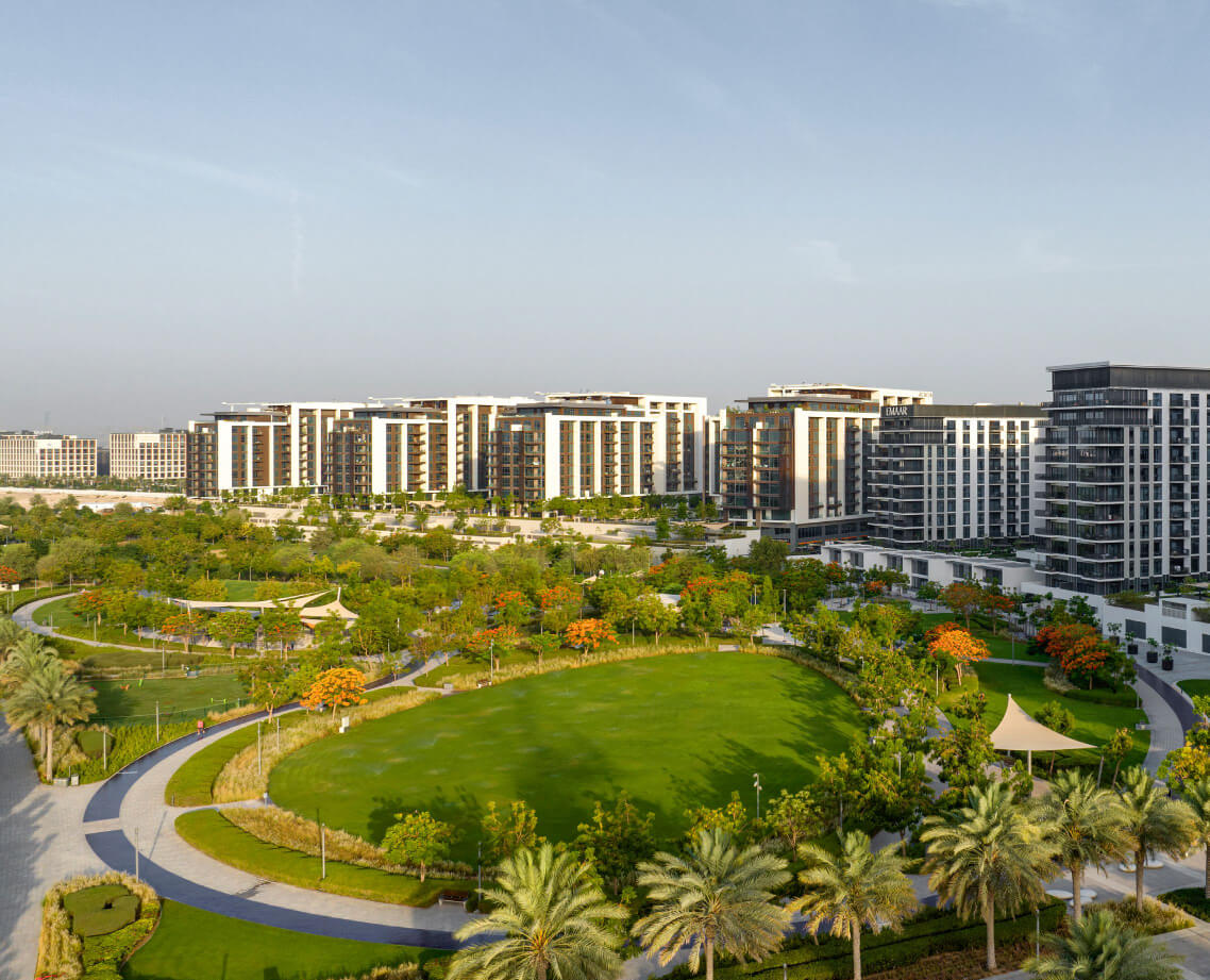 A landscaped urban park with green lawns, palm trees, walking paths, and shaded seating areas, surrounded by modern mid-rise Flats in Dubai under a clear sky.